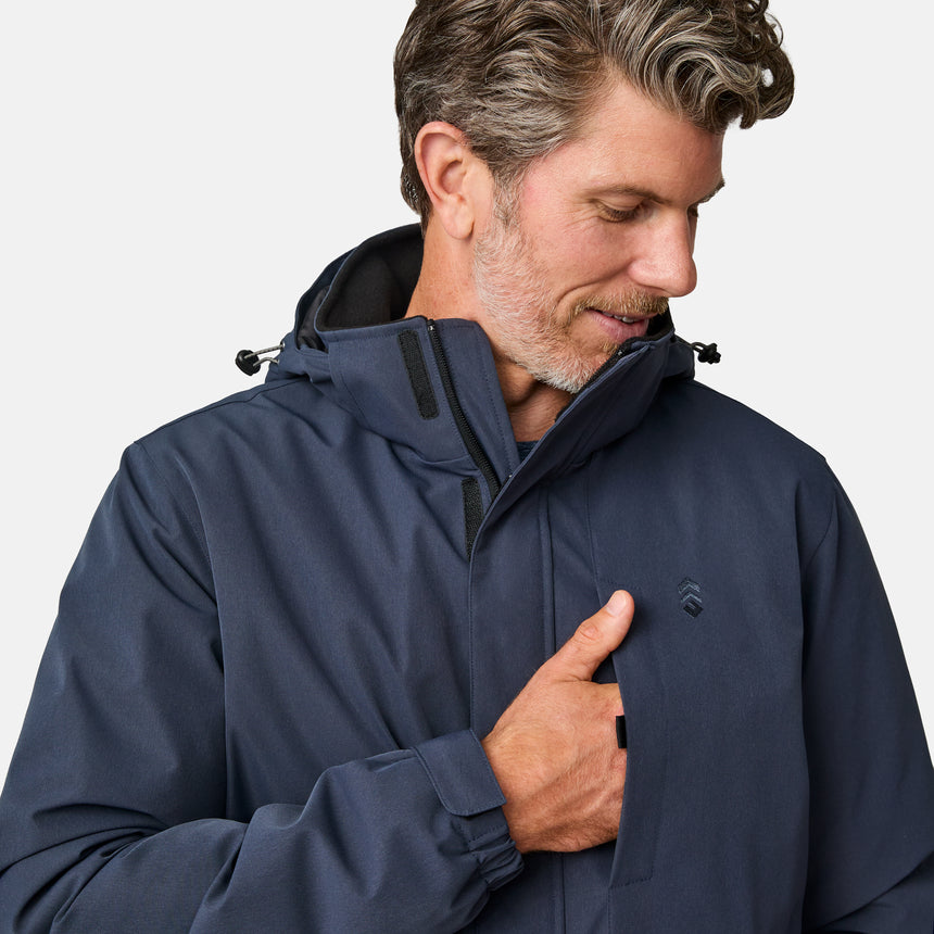[Kolby is 6’2” wearing size M.] Men’s Free Country navy zip-front jacket with hood and high collar, featuring vertical chest zipper pocket and side hand pockets—worn over light brown pants for an outdoors-ready, insulated look.