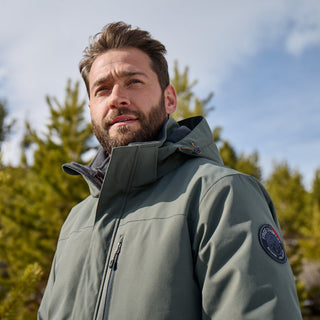 A bearded man wears the Free Country Men's Ski Patrol 3-in-1 Systems Jacket in green, standing outdoors before pine trees, looking upward as sunlight brightens his face beneath a partly cloudy sky.