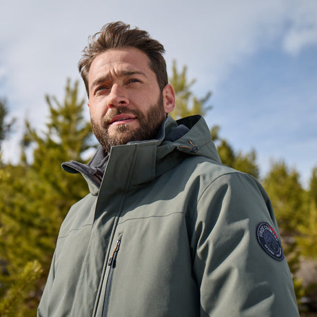 A bearded man wears the Free Country Men's Ski Patrol 3-in-1 Systems Jacket in green, standing outdoors before pine trees, looking upward as sunlight brightens his face beneath a partly cloudy sky.