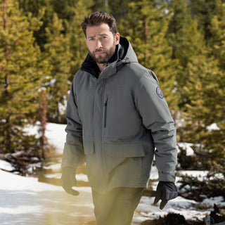 A bearded man wearing the Free Country Men's Ski Patrol 3-in-1 Systems Jacket stands in a snowy pine forest, black gloves on, looking at the camera with a neutral expression.