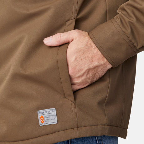 [Joe is 6'0" wearing size M.] Men’s Free Country brown canvas jacket layered over a white crewneck tee and blue jeans—features zippered chest pockets and left-side logo patch for rugged outdoor appeal and everyday versatility.