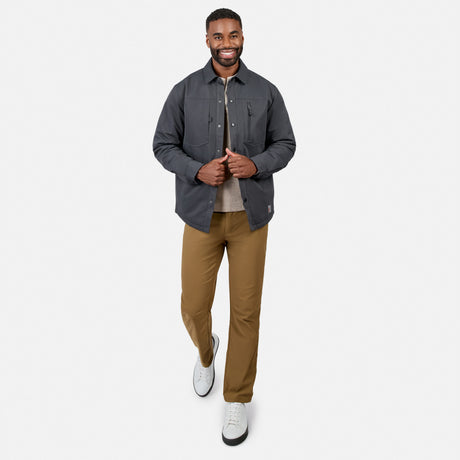 A man smiles while walking forward, dressed in the Free Country Men’s Brawny Canvas Function Work Jacket—a dark gray water-resistant piece—paired with a beige shirt, tan pants, and white sneakers against a plain white background.