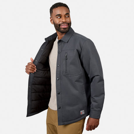 A man smiles while modeling the Free Country Men’s Brawny Canvas Function Work Jacket in dark gray, featuring snap buttons and a chest pocket. He holds one side open to reveal the quilted lining of this eco-conscious jacket, which is styled with a beige shirt and tan pants against a plain white background.