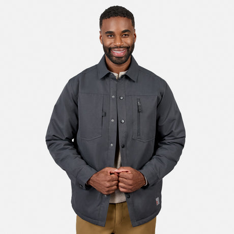 Smiling man wearing the Free Country Men’s Brawny Canvas Function Work Jacket in dark gray over a light sweater and brown pants, standing against a plain white background.