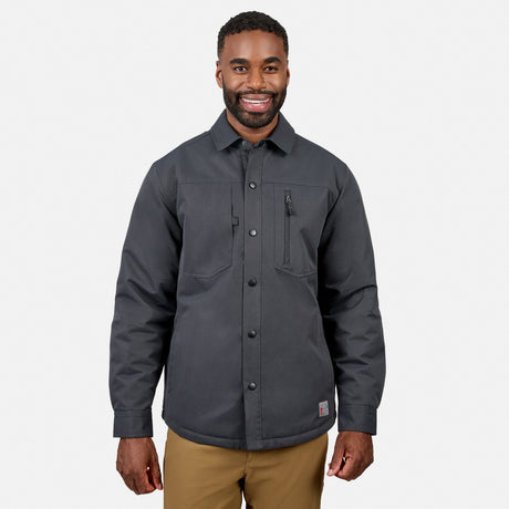 A man stands smiling against a plain white background wearing the Free Country Men’s Brawny Canvas Function Work Jacket in dark gray, featuring two chest pockets, along with tan pants.
