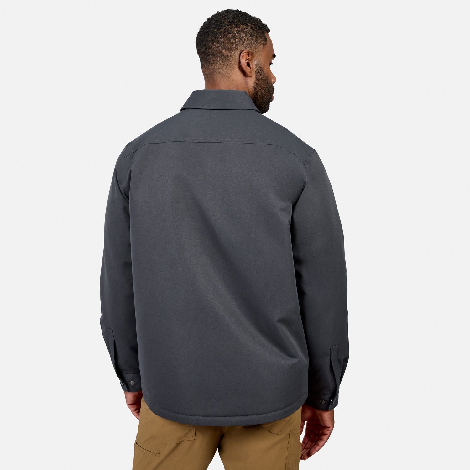 A man stands facing away, wearing the Free Country Men’s Brawny Canvas Function Work Jacket in dark gray and tan pants against a plain white background.