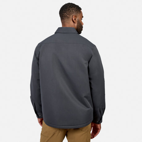 A man stands facing away, wearing the Free Country Men’s Brawny Canvas Function Work Jacket in dark gray and tan pants against a plain white background.