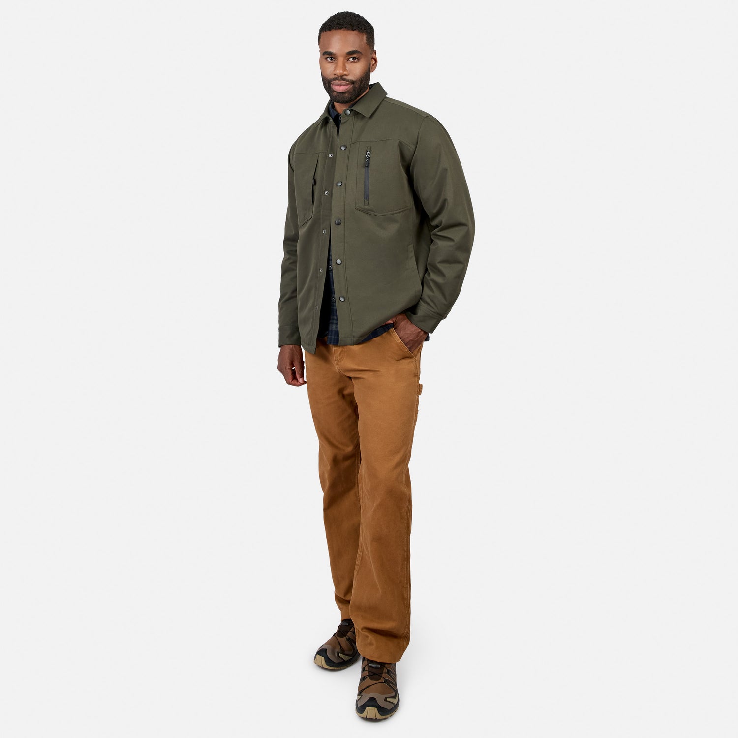 A man stands against a white background wearing the Men’s Brawny Canvas Function Work Jacket by Free Country in olive green, paired with a black shirt, brown pants, and camouflage sneakers, one hand in his pocket.