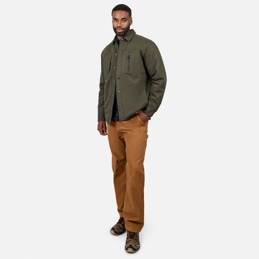 A man stands against a white background wearing the Men’s Brawny Canvas Function Work Jacket by Free Country in olive green, paired with a black shirt, brown pants, and camouflage sneakers, one hand in his pocket.