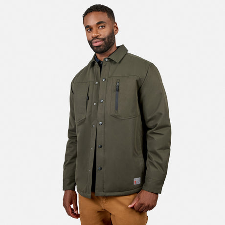 A man wearing the Free Country Men’s Brawny Canvas Function Work Jacket in olive green, paired with tan pants, stands against a plain white background and looks at the camera with a slight smile.
