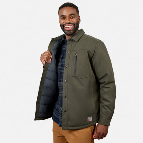 A smiling bearded man models the Free Country Men’s Brawny Canvas Function Work Jacket in dark olive green over a plaid shirt and tan pants, holding open one side to reveal the jacket’s black insulated lining, set against a plain light background.