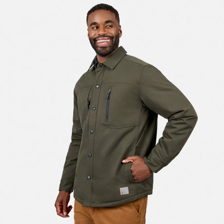 A man is smiling and standing against a plain white background, wearing the Free Country Men’s Brawny Canvas Function Work Jacket in olive green with chest pockets, paired with brown pants.