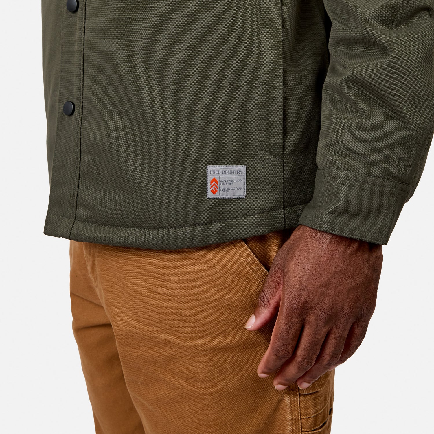 A person is wearing the Men’s Brawny Canvas Function Work Jacket by Free Country in olive green, paired with light brown pants. The water-resistant jacket features a visible label, and the image highlights their lower torso and right hand against a plain background.