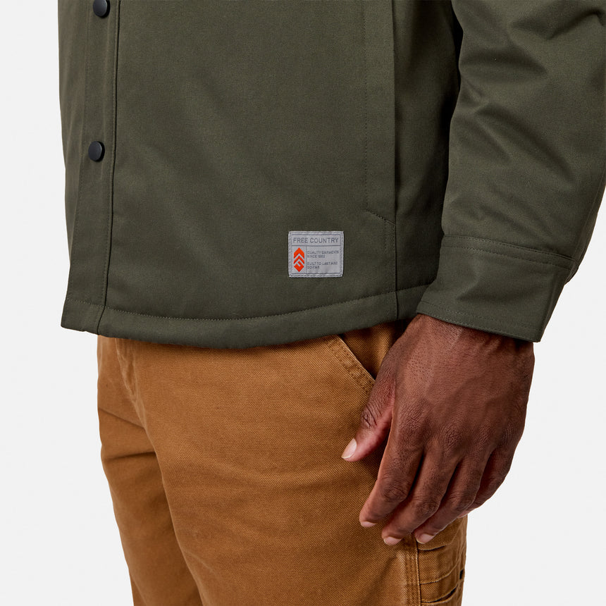 A person is wearing the Men’s Brawny Canvas Function Work Jacket by Free Country in olive green, paired with light brown pants. The water-resistant jacket features a visible label, and the image highlights their lower torso and right hand against a plain background.