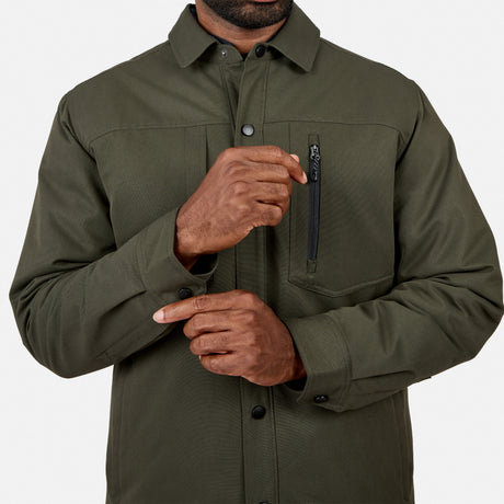 A person wearing the Free Country Men’s Brawny Canvas Function Work Jacket in olive green, featuring a zippered chest pocket and eco-conscious design, adjusts the cuff on their left sleeve. Their face is not visible.