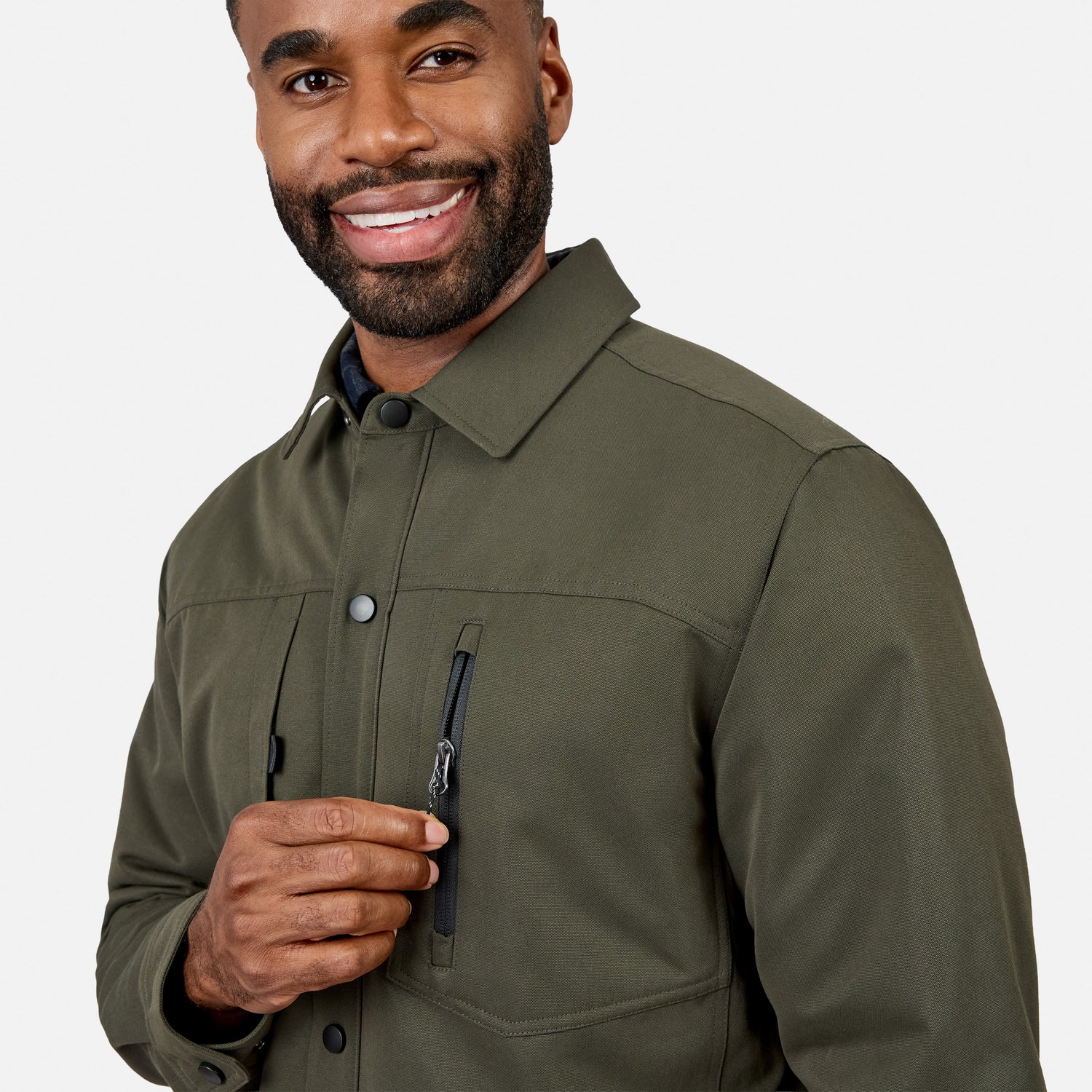 A man wearing the Free Country Men’s Brawny Canvas Function Work Jacket in olive green smiles while holding the zipper of a chest pocket. The background is plain white.