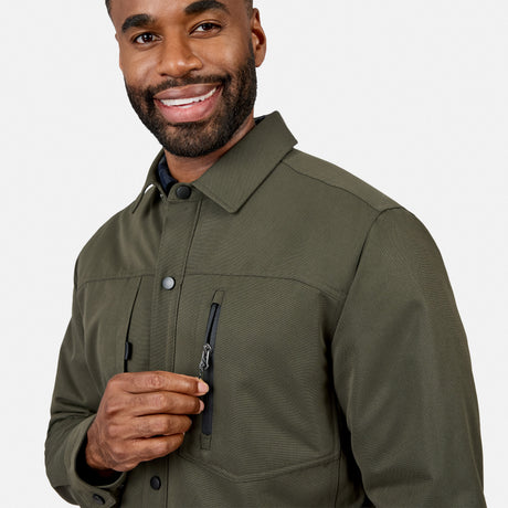 A man wearing the Free Country Men’s Brawny Canvas Function Work Jacket in olive green smiles while holding the zipper of a chest pocket. The background is plain white.