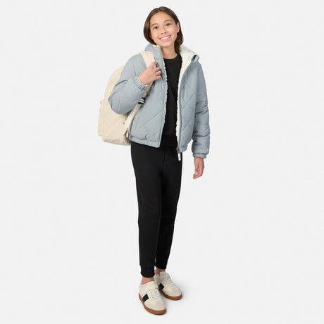 A young girl stands against a light gray studio background wearing a light blue puffer jacket with a cream sherpa lining, black pants, and beige sneakers, while carrying a cream backpack.