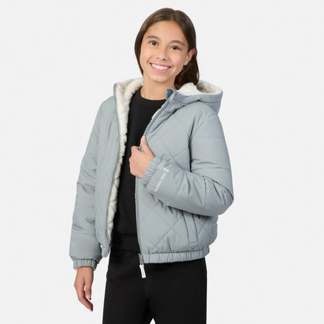 A young girl smiling and wearing a light gray hooded puffer jacket with a white sherpa lining, paired with a black shirt and black pants, against a light gray studio background.