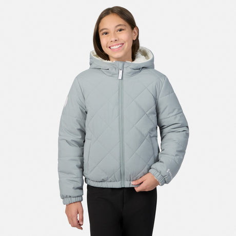 A young girl smiling while wearing a light gray diamond-quilted hooded puffer jacket with a white sherpa lining and black pants, set against a plain light gray background.