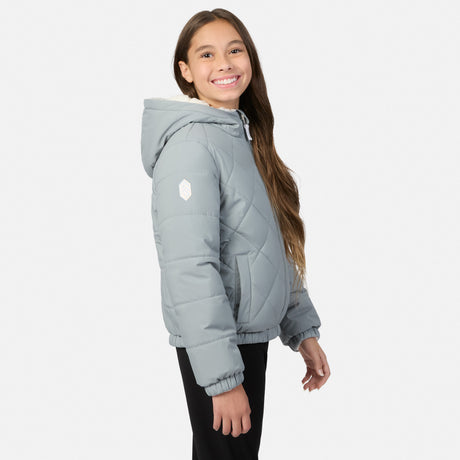 A smiling young girl with long brown hair models a light gray quilted puffer jacket with a white geometric logo on the sleeve, paired with black pants, against a solid off-white background.