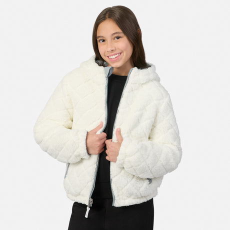 A young girl smiling while wearing a cream-colored, quilted sherpa jacket with a gray zipper, styled over a black top and black pants, against a light gray studio background.