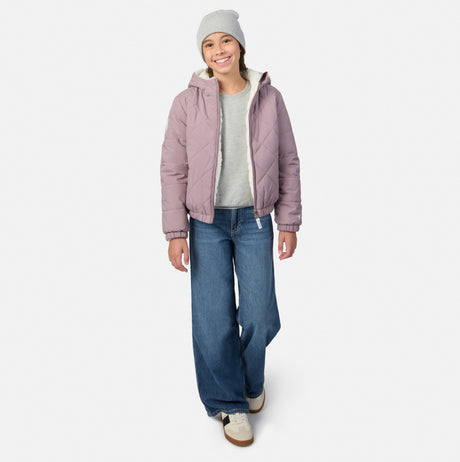 A woman modeling a light purple quilted puffer jacket with a hood, worn over a light gray t-shirt and blue wide-leg jeans, paired with a light gray beanie and white sneakers, on a light gray background.
