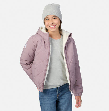 A young girl smiling while wearing a dusty purple quilted puffer jacket with a white sherpa lining, a light gray beanie, a gray t-shirt, and blue jeans, set against a plain light gray background.
