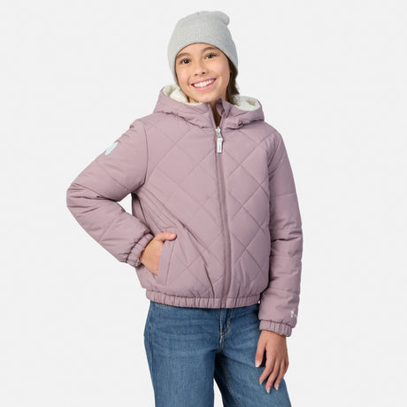 A young girl wearing a dusty purple quilted hooded puffer jacket with a white fleece lining, blue jeans, and a light gray beanie, standing against a light gray studio background.