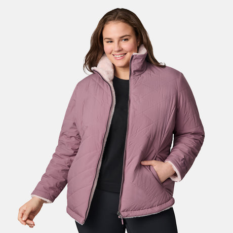 A woman with long brown hair models the Clearance Women's Plus Size Cirrus Quilted Reversible Jacket in mauve with a light pink fleece lining, over a black shirt and pants, posing against a plain white background and smiling slightly.