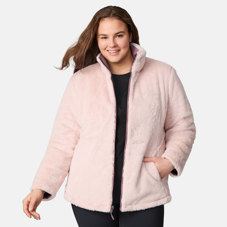 A woman smiles while wearing the Clearance Women's Plus Size Cirrus Quilted Reversible Jacket, made with recycled polyester, over a black top and pants. She has one hand in her pocket and stands against a light gray background.