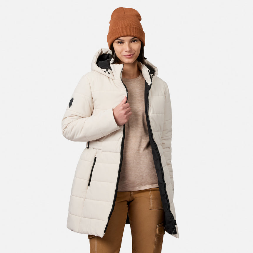 A woman wears a cream-colored, long quilted hooded Women’s Cirrus Quilted Long Puffer Jacket by Free Country over a tan shirt and brown pants, finished with a rust-colored beanie.