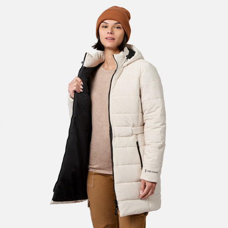 A person wearing an off-white, long quilted Women’s Cirrus Quilted Long Puffer Jacket from Free Country with REPREVE insulation and black lining, paired with a tan beanie and tan pants.