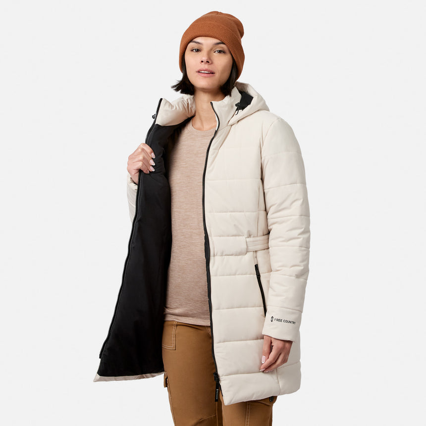 A person wearing an off-white, long quilted Women’s Cirrus Quilted Long Puffer Jacket from Free Country with REPREVE insulation and black lining, paired with a tan beanie and tan pants.