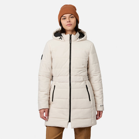 A woman wearing a bisque Free Country Women’s Cirrus Quilted Long Puffer Jacket featuring a hood, black zippers, and a waist belt.