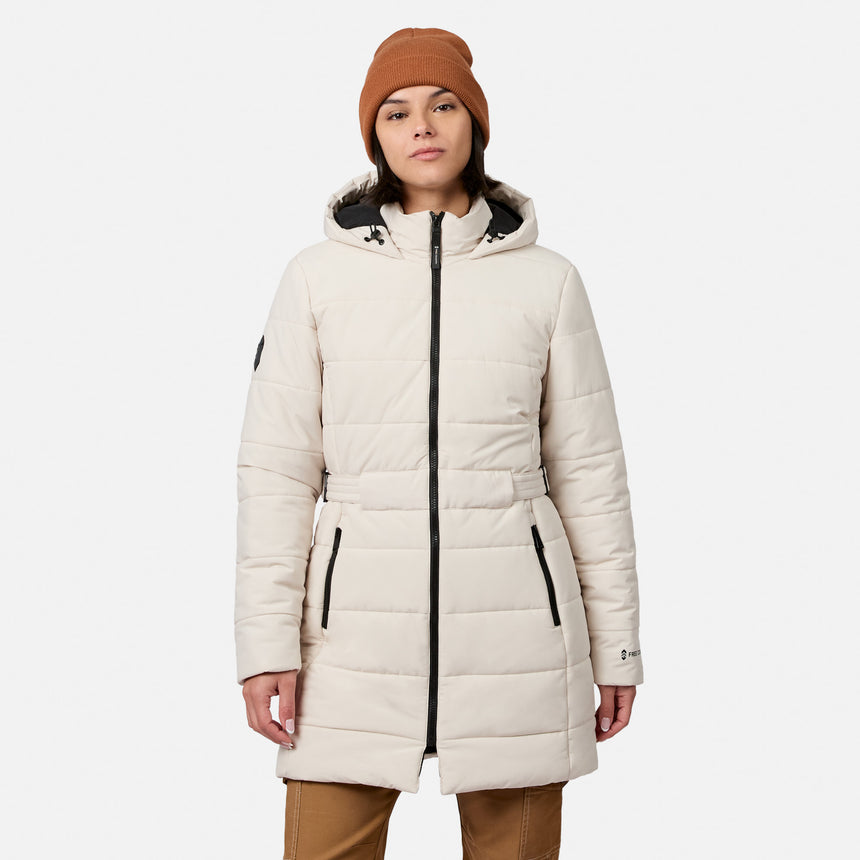 A woman wearing a bisque Free Country Women’s Cirrus Quilted Long Puffer Jacket featuring a hood, black zippers, and a waist belt.