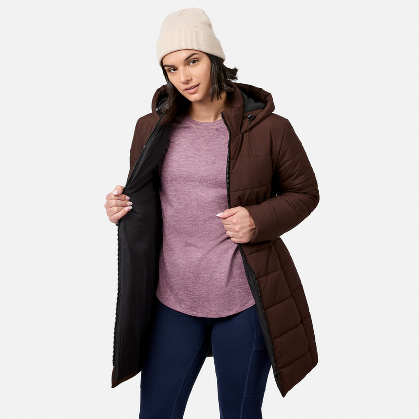A person wearing a beige beanie and a dark brown Free Country Women’s Cirrus Quilted Long Puffer Jacket open over a purple t-shirt and dark leggings.