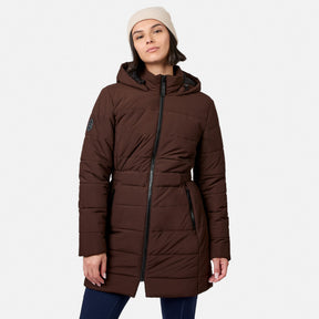 A woman wearing an espresso-colored Women’s Cirrus Quilted Long Puffer Jacket, a dark brown recycled polyester hooded coat with a front zipper and belted waist.