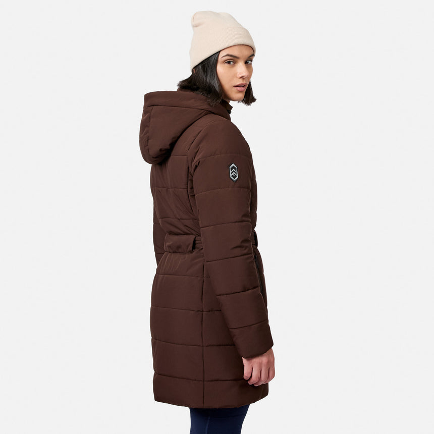 A woman wearing a brown Free Country Women’s Cirrus Quilted Long Puffer Jacket with recycled insulation and a beige beanie looking over her shoulder.