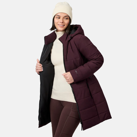 A woman wears a burgundy, water-resistant Free Country Women’s Cirrus Quilted Long Puffer Jacket with a hood, a cream turtleneck sweater, and a matching beanie.