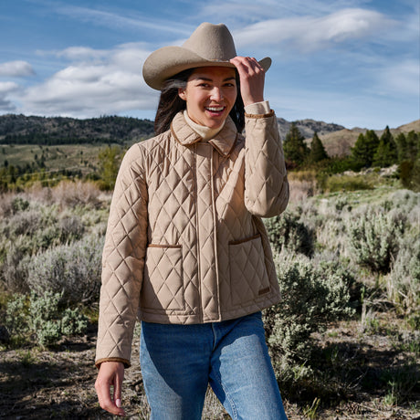 A woman wearing a beige quilted jacket, blue jeans, and a wide-brim cowboy hat stands outdoors in a grassy, mountainous landscape under a blue sky.