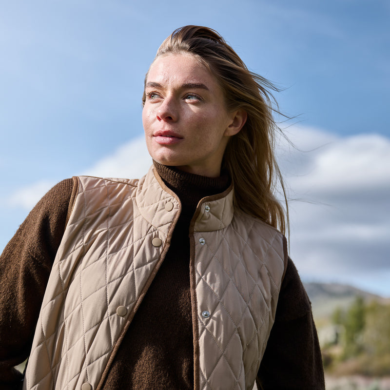 A woman wearing the Free Country Women’s Cirrus Diamond Quilted Button Down Vest in beige, layered over a brown sweater, stands outdoors gazing into the distance. The backdrop features a blue sky with soft clouds, along with touches of greenery and distant mountains.