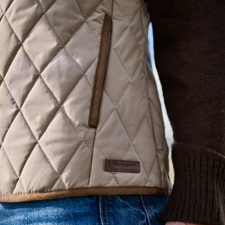 A close-up of a person wearing the Women’s Cirrus Diamond Quilted Button Down Vest by Free Country—a beige diamond quilted vest with brown trim, styled over a dark brown sweater and blue jeans. A small label on the recycled polyester vest reads TREE COUNTRY.