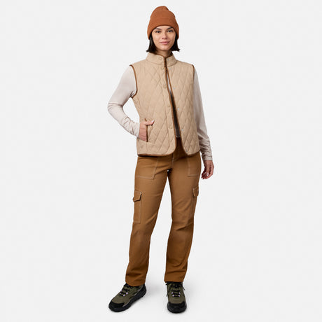 A woman stands against a plain white background, smiling with one hand in her pocket, wearing a brown beanie, long-sleeve shirt, brown cargo pants, black hiking shoes, and the Free Country Women’s Cirrus Diamond Quilted Button Down Vest.