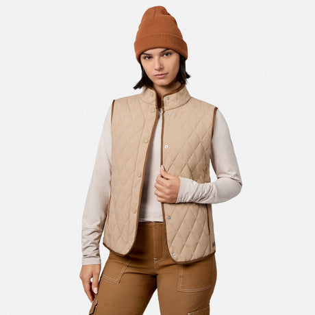 A woman wearing the Free Country Women’s Cirrus Diamond Quilted Button Down Vest in almond over a long-sleeve shirt, paired with brown pants and a brown knit beanie, stands against a plain white background.