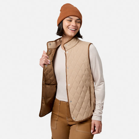 A woman smiles with her eyes closed against a plain light background, wearing the Free Country Women’s Cirrus Diamond Quilted Button Down Vest in tan over a light long sleeve shirt, paired with brown pants and a brown beanie.