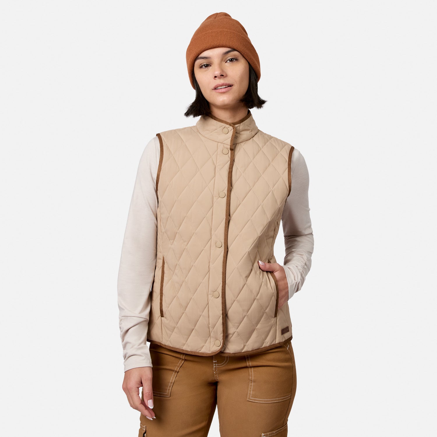A person wearing the Free Country Women’s Cirrus Diamond Quilted Button Down Vest in tan over a beige long-sleeve shirt, brown pants, and a brown beanie stands with one hand in their vest pocket against a plain white background.