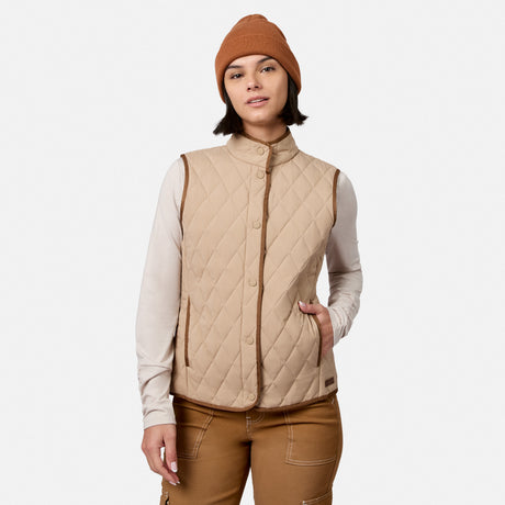 A person wearing the Free Country Women’s Cirrus Diamond Quilted Button Down Vest in tan over a beige long-sleeve shirt, brown pants, and a brown beanie stands with one hand in their vest pocket against a plain white background.