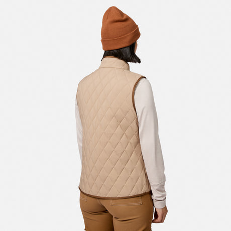 A woman is shown from the back against a plain white background wearing the Free Country Women’s Cirrus Diamond Quilted Button Down Vest in tan, styled with a light long-sleeve shirt, brown pants, and a brown beanie.