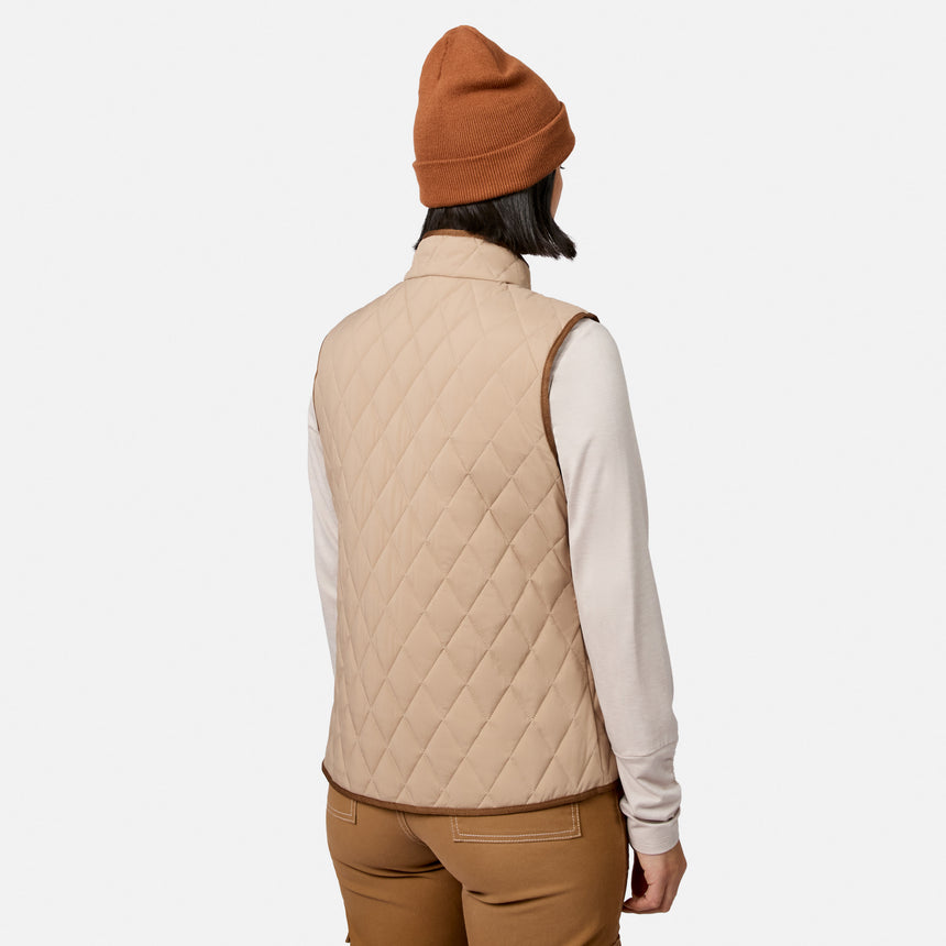 A woman is shown from the back against a plain white background wearing the Free Country Women’s Cirrus Diamond Quilted Button Down Vest in tan, styled with a light long-sleeve shirt, brown pants, and a brown beanie.